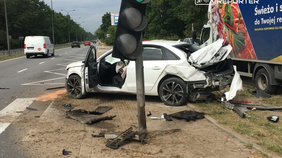 Rozbity biały samochód osobowy stoi przy sygnalizatorze, z uszkodzeniami z tyłu i otwartymi drzwiami, na poboczu drogi.