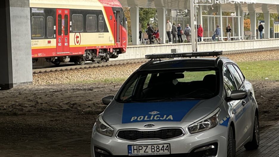 Radiowóz policyjny zaparkowany na stacji kolejowej, obok przejeżdżającego pociągu. W tle widoczni pasażerowie czekający na peronie.