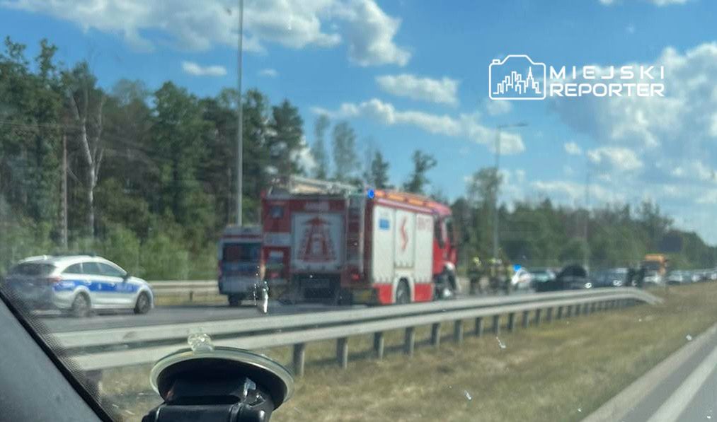 Na drodze widoczny wóz strażacki oraz radiowóz policyjny, w tle zablokowany ruch pojazdów.