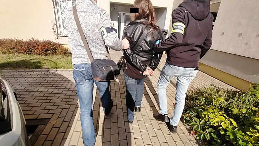Kobieta w skórzanej kurtce prowadzona przez dwóch policjantów na chodniku przed budynkiem.