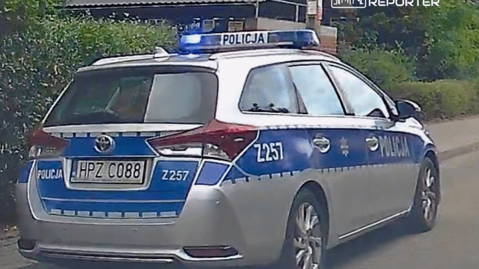 Policyjny radiowóz z niebieskimi światłami zaparkowany na ulicy, otoczony zielenią.