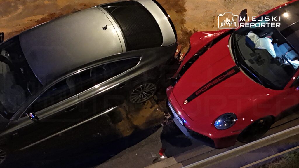 Dwa zaparkowane samochody, srebrny sedan i czerwone Porsche, zderzone ze sobą na ulicy nocą.
