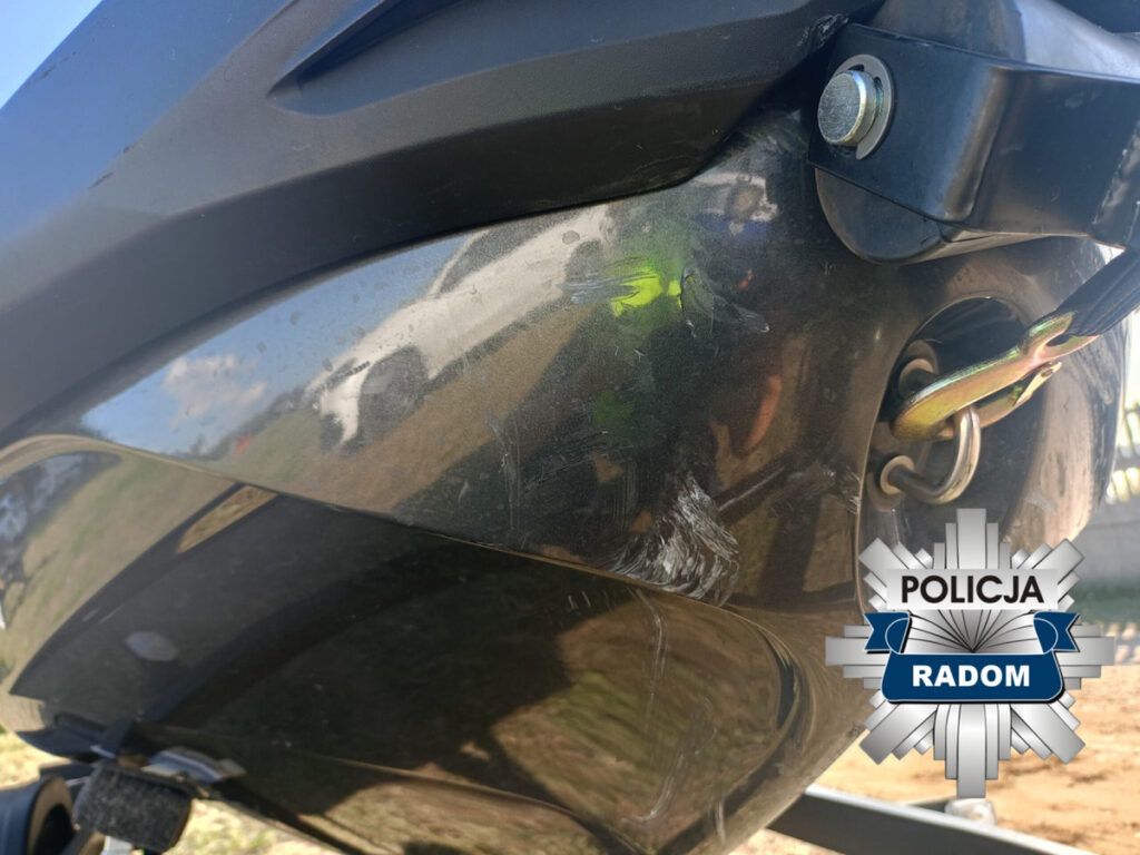 Zarysowana tylna część motocykla zaparkowanego na trawie, w tle widoczny jasny samochód.