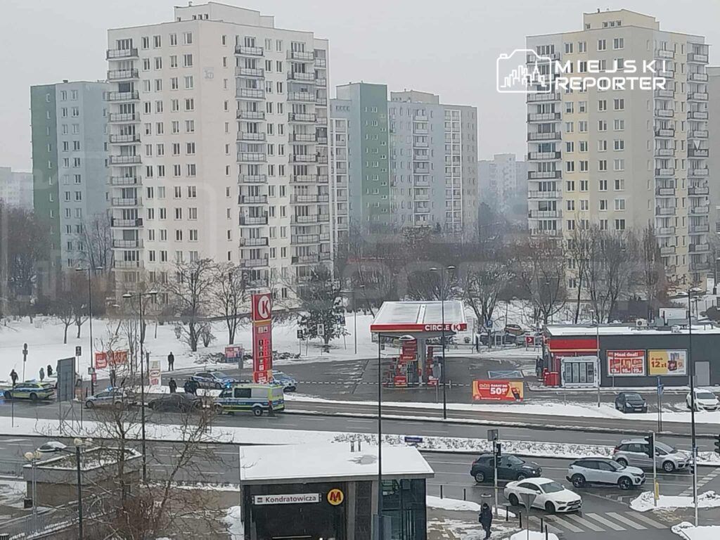 Grupa ludzi obserwująca interwencję policji przy stacji benzynowej w zimowym otoczeniu, w tle bloki mieszkalne.