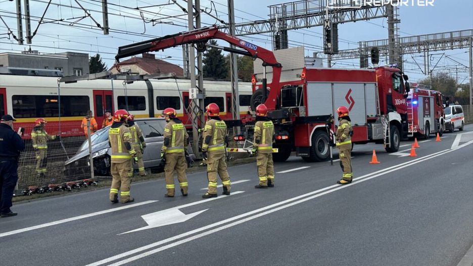 Grupa strażaków w mundurach z hełmami pracuje przy uszkodzonym samochodzie obok torów kolejowych.
