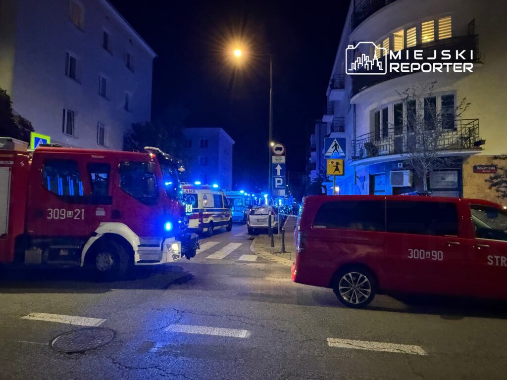 Czerwony wóz strażacki z włączonymi sygnalizatorami na oświetlonej ulicy, obok ambulansy i policyjne pojazdy.