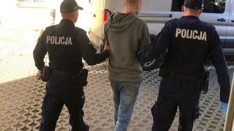 Dwóch policjantów prowadzi zatrzymanego mężczyznę w szarej bluzie na podwórzu.