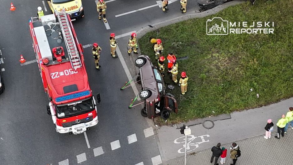 Wielu strażaków pracuje przy przewróconym samochodzie na poboczu drogi, obok straży pożarnej i ambulansu.