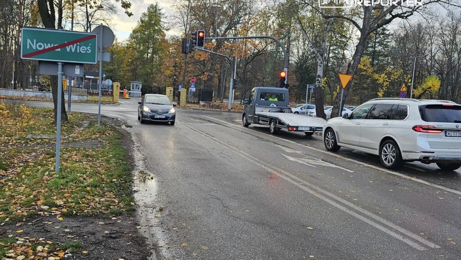 Ruch samochodowy na skrzyżowaniu z sygnalizacją świetlną, w tle znak drogowy "Nowa Wieś". Jesienna sceneria z opadającymi liśćmi.