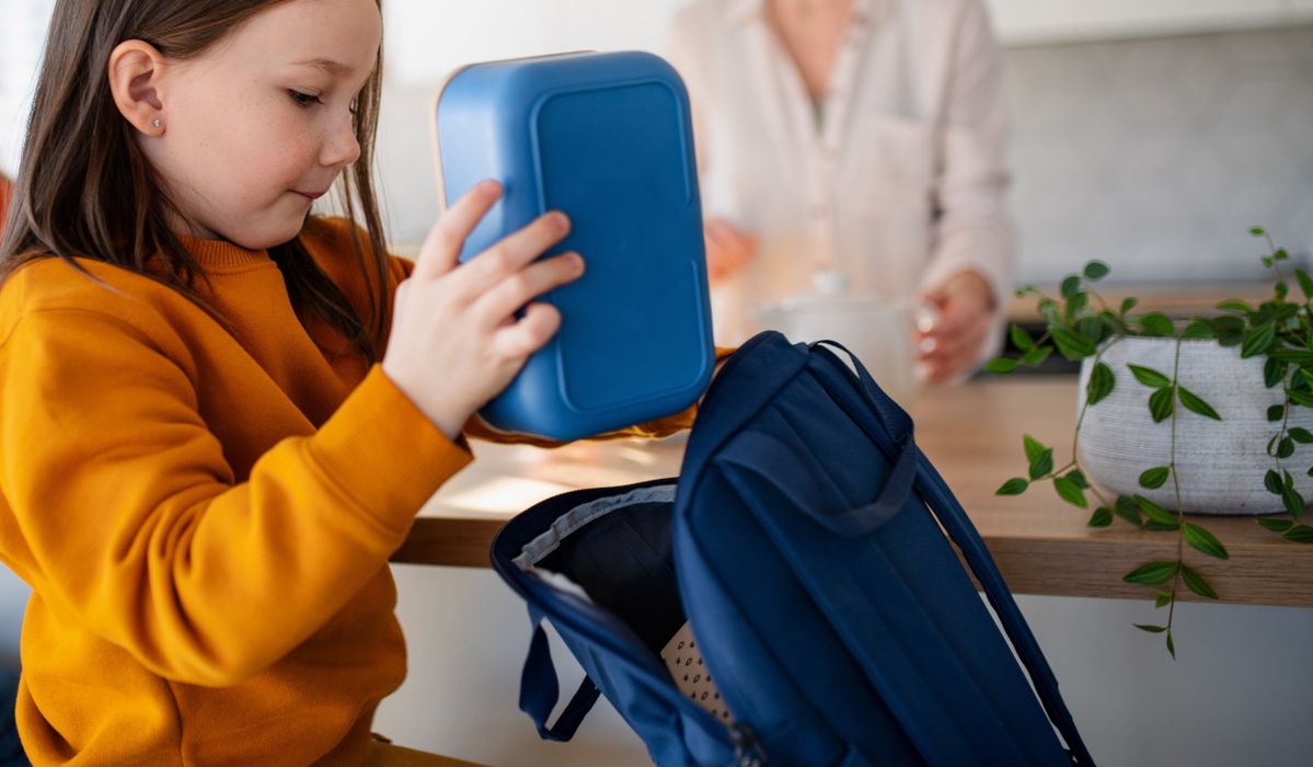 A little girl packing lunch box to backpack in kitchen at home.