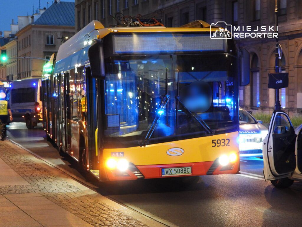 Autobus miejski zatrzymany na ulicy, obok radiowóz policyjny z włączonymi sygnałami świetlnymi.