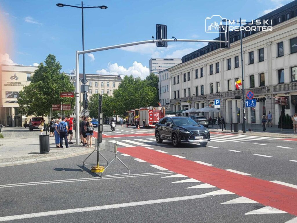 Grupa ludzi stojąca na chodniku obok czerwonej strefy na ulicy, przejeżdżający czarny samochód oraz w tle wozów strażackich.