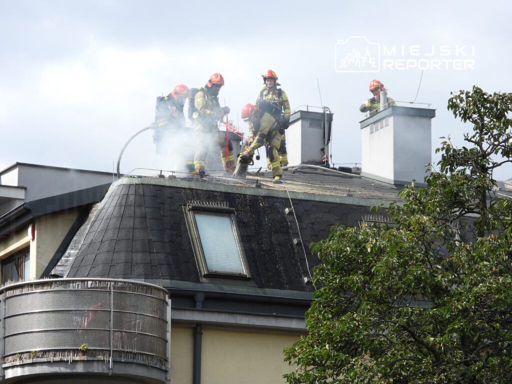 Grupa strażaków w hełmach gasi pożar na dachu budynku, z którego unosi się dym.