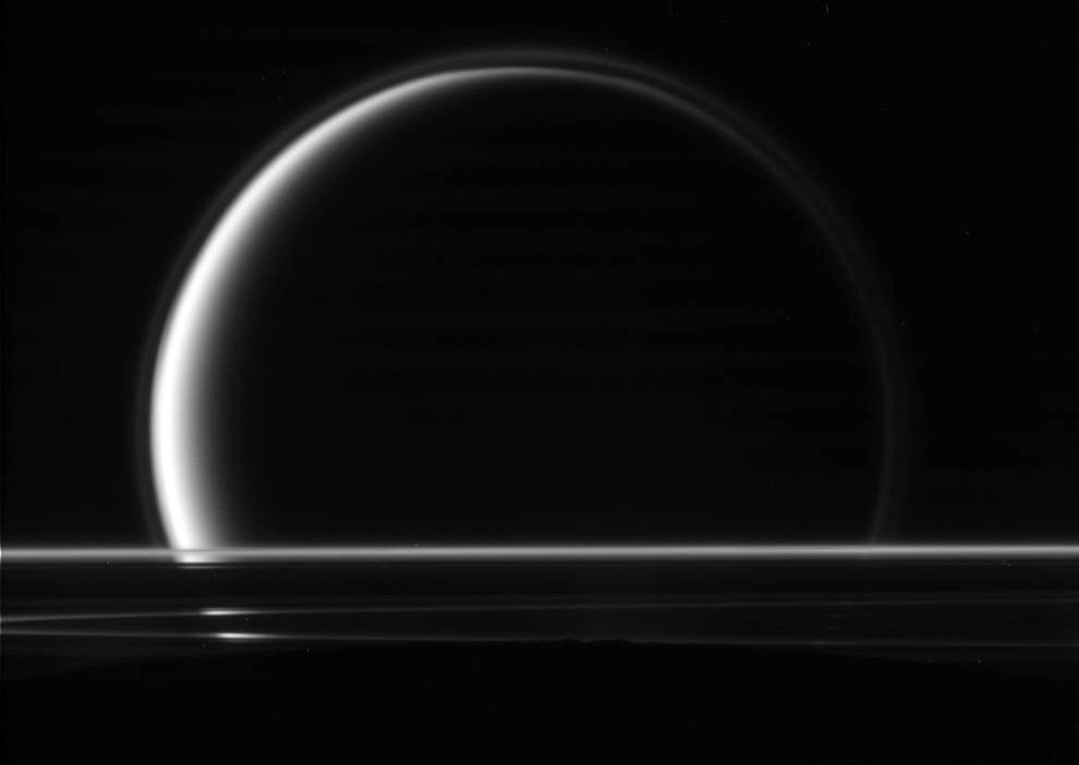 Spowity w gęstą, azotowo-metanową atmosferę Tytan, widziany ponad krawędzią lodowo-skalistego Enceladusa (czarny obszar na dole fotografii). Pasy pomiędzy dwoma satelitami stanowią fragment pierścieni Saturna. Zdjęcie wykonano 18 maja 2010 r.