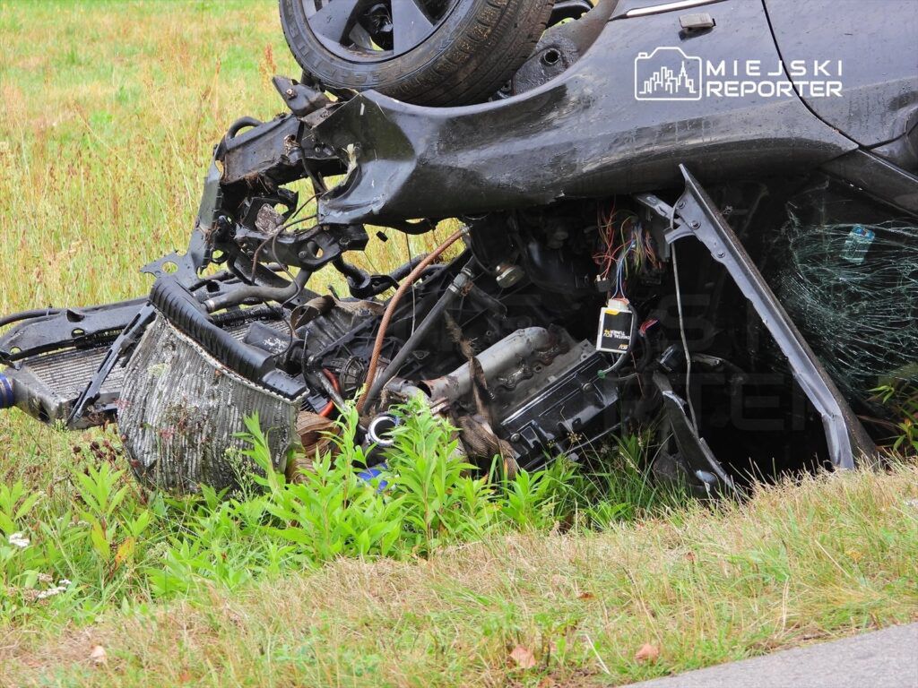 Zniszczony samochód leży na boku w wysokiej trawie, odsłaniając uszkodzone podzespoły silnika.