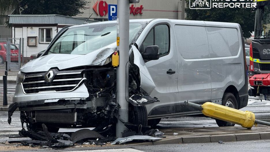 Uszkodzony biały furgon Renault uderzył w słup sygnalizacji świetlnej na deszczowej ulicy, obok sklepu.