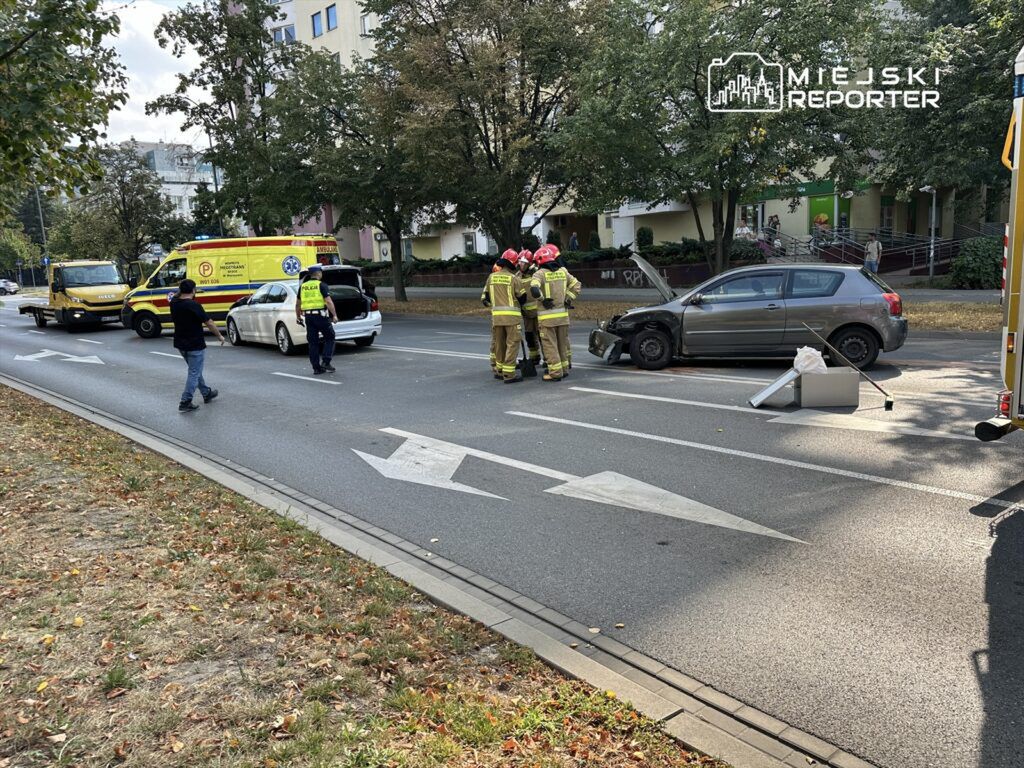 Grupa strażaków w hełmach omawia sytuację przy uszkodzonym samochodzie na ulicy, obok parkującego ambulansu.