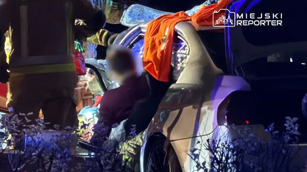 Ratownicy medyczni pomagają osobie wydobytej z rozbitego samochodu w nocy, otoczeni przez pojazdy służb ratunkowych.