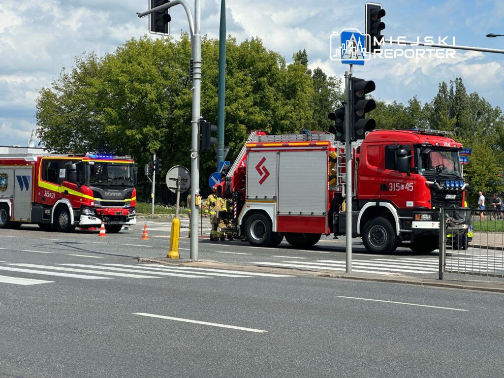Na skrzyżowaniu ulicy widać dwa wozy strażackie z włączonymi sygnałami świetlnymi. Strażacy pracują przy jednym z pojazdów, a na drodze ustawione są pachołki ostrzegawcze. W tle widać zieleń oraz sygnalizację świetlną. Sytuacja może być związana z akcją ratunkową lub kontrolą drogową.