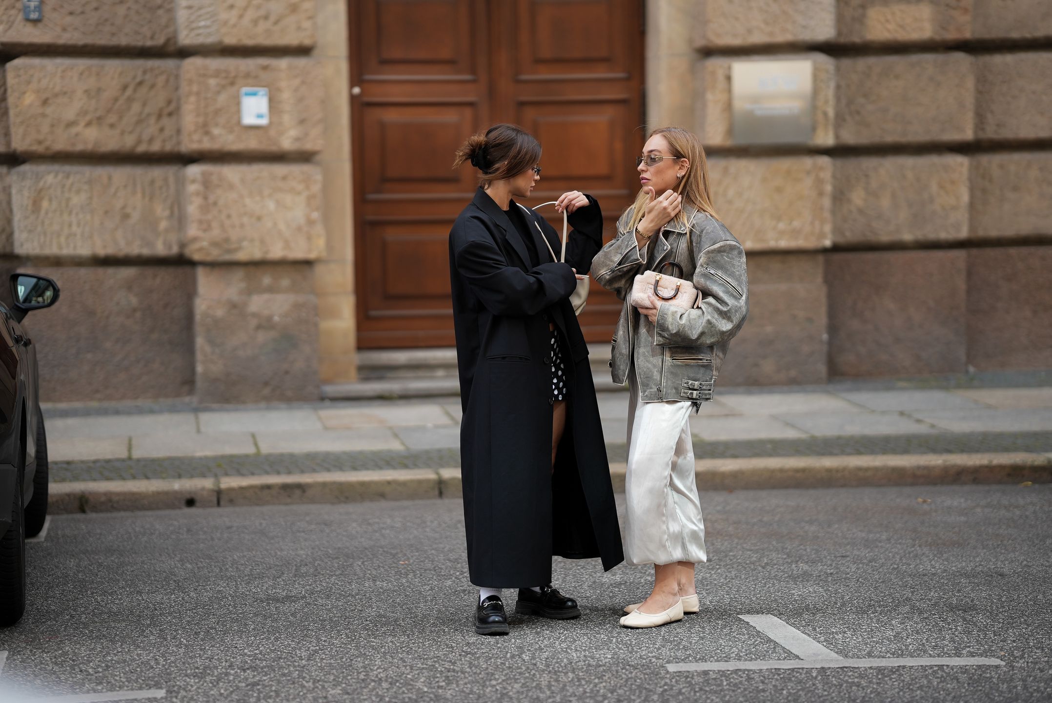 BERLIN, GERMANY - SEPTEMBER 1: Sonia Lyson (right) is seen wearing a black lace camisole with delicate trim from Zara; an oversized grey leather biker jacket with a distressed finish and zipper details from Ducie; Wide-leg satin trousers in an off-white shade with a voluminous, flowy silhouette from Zara; beige ballet flats with a minimal, classic shape from Margiela; a soft blush pink pouch bag with embroidered branding and gold details from Fendi; slim, see through, oval-shaped sunglasses with slightly dark tintedlenses and golden fram from MiuMiu; her blonde hair is worn down with a side part; Sophia Geiss (left) is seen wearing a long, oversized black coat with broad shoulders and a straight cut, worn open from 032C; a simple black crop top, leaving the midriff slightly visible from Zara; a short, fitted black-and-white polka dot mini skirt from Attega; black chunky loafers paired with white socks from LeGer by Lena Gercke; a medium-sized white handbag with shoulder straps from Demellier London; small rectangular black sunglasses with a modern touch from Vela Eyewear; her brown hair is pulled back into a slightly messy low bun on September 1, 2025 in Berlin, Germany. (Photo by Moritz Scholz/Getty Images)