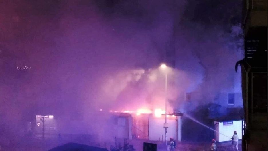 Ognisko pożaru otoczone gęstym dymem, w tle strażacy gaszący płomienie w nocy.