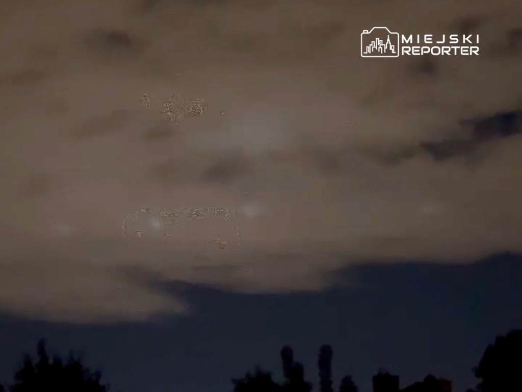 Nocne niebo z chmurami, w tle widoczne słabe punkty światła, otoczenie z siluetami drzew.