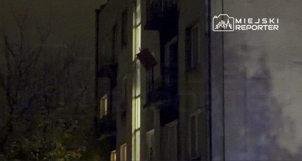 Czerwony przedmiot zwisający z balkonu na drugim piętrze budynku mieszkalnego w ciemnej scenerii nocnej.