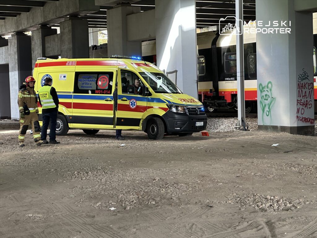 Zespół ratownictwa medycznego rozmawia z mężczyzną obok karetki pod wiaduktem kolejowym, w tle pociąg.
