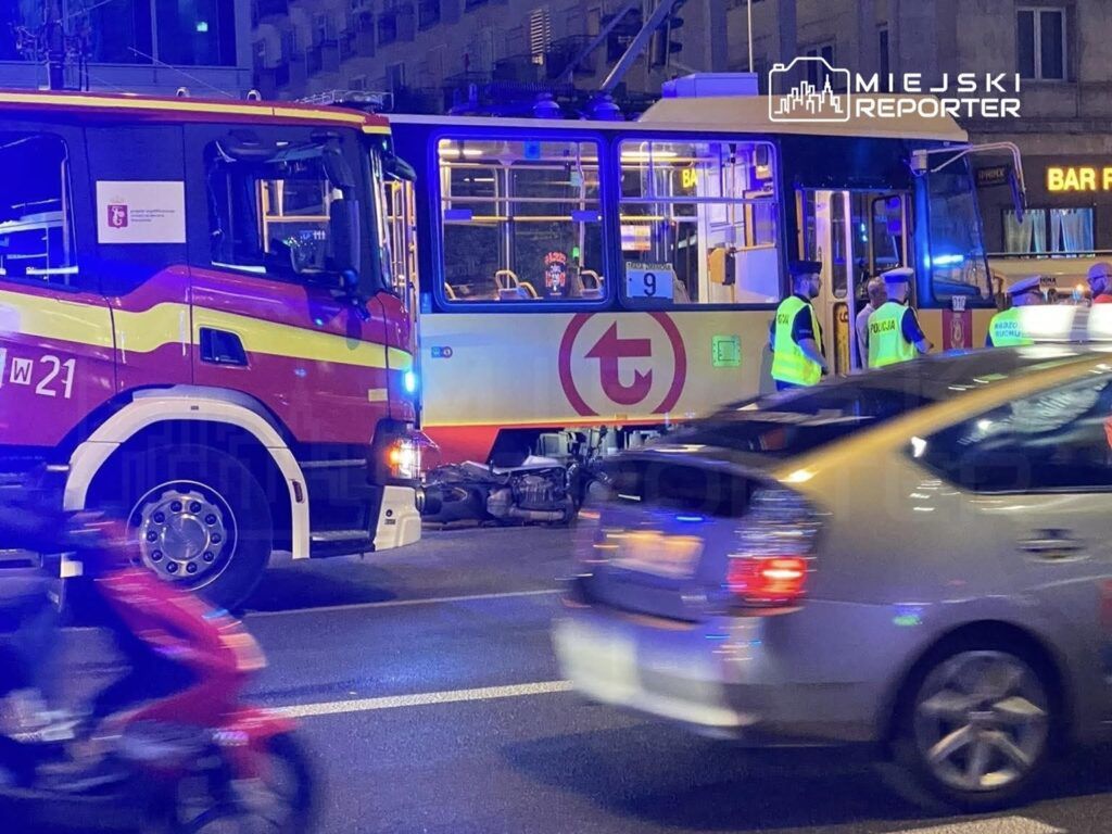 Pojazd strażacki oraz policjanci w kamizelkach odblaskowych zabezpieczają miejsce wypadku tramwaju w ruchliwej ulicy.