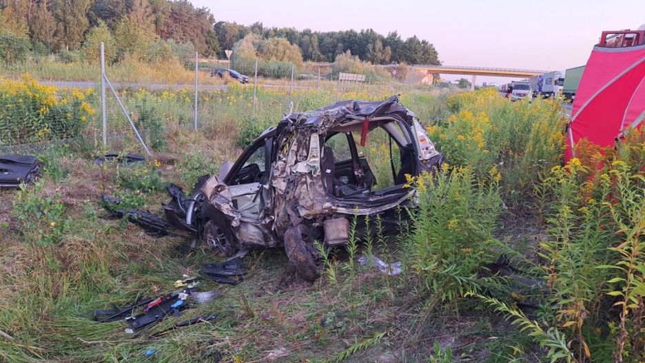 Zniszczony samochód leży w trawie obok drogi, otoczony dziką roślinnością i ogrodzeniem.