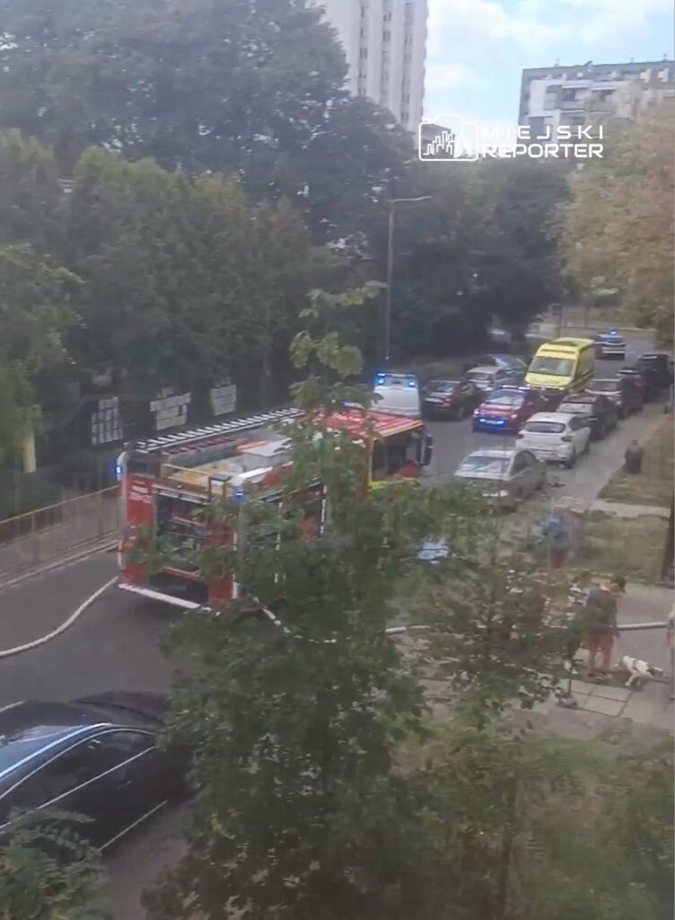 Czerwony wóz strażacki z włączonymi sygnałami świetlnymi stoi na ulicy, obok zaparkowane samochody i karetka.