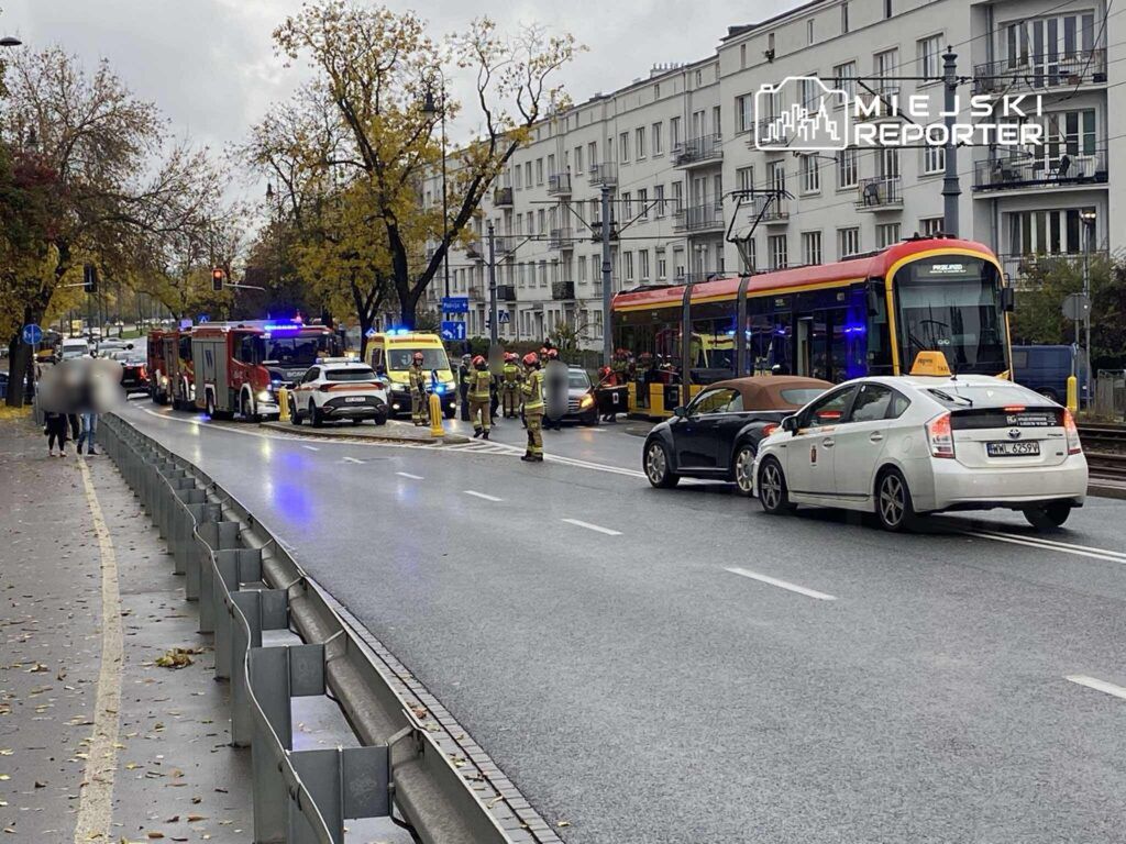 Na ulicy, w deszczowej aurze, strażacy oraz policja pracują przy wypadku drogowym, obok zatrzymany tramwaj.