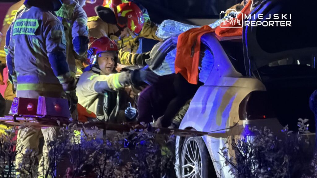 Grupa strażaków w hełmach i mundurach, pomagająca w wydobywaniu osoby z wraku samochodu w ciemnej scenerii.