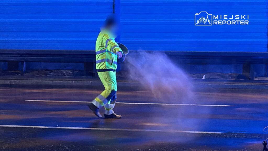 Pracownik w żółtej odzieży odblaskowej rozsypuje piasek na mokrej nawierzchni drogi w nocy.