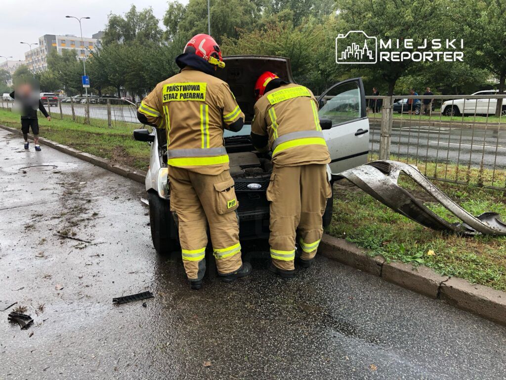Dwaj strażacy w mundurach przeglądają silnik samochodu osobowego na poboczu drogi, obok leżą fragmenty płyty.