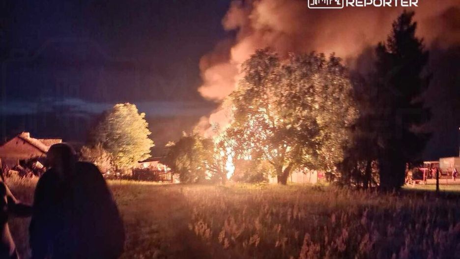 Ognisko pożaru o jasnych płomieniach w pobliżu drzew, w tle widoczne zarysy budynków i dym unoszący się w nocy.