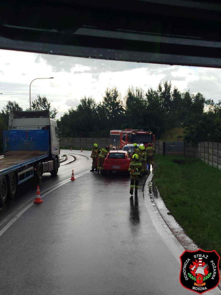 Grupa strażaków w mundurach omawia sytuację przy czerwonym samochodzie na mokrej drodze, obok ciężarówki.