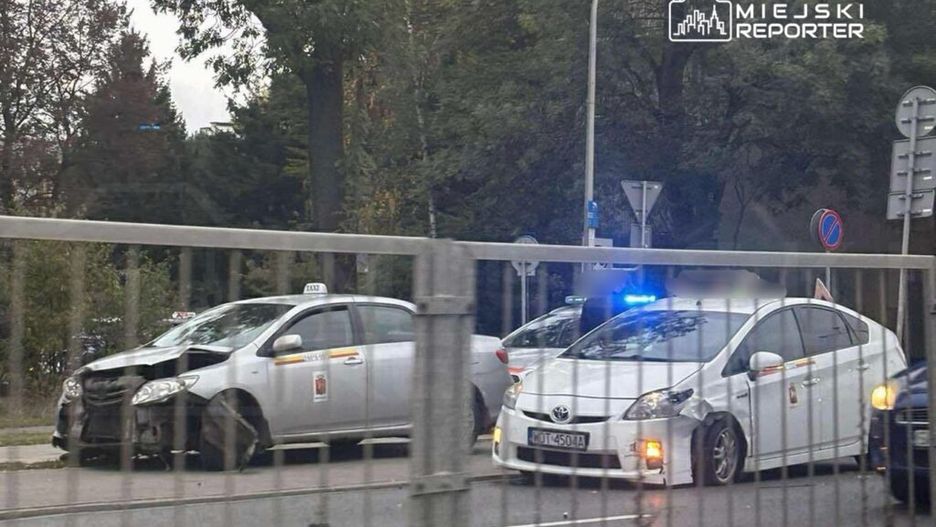 Dwa uszkodzone taksówki stoją na poboczu drogi, obok policyjny radiowóz z włączonymi sygnałami.