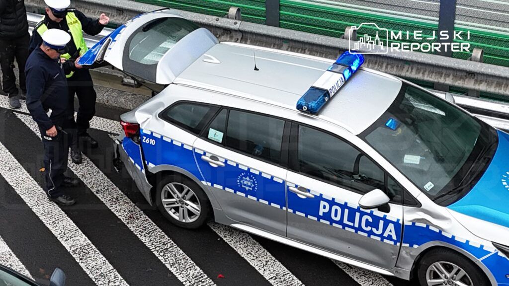 Dwaj policjanci sprawdzają uszkodzenie radiowozu na pasie drogowym, obok widoczna bariera ochronna.
