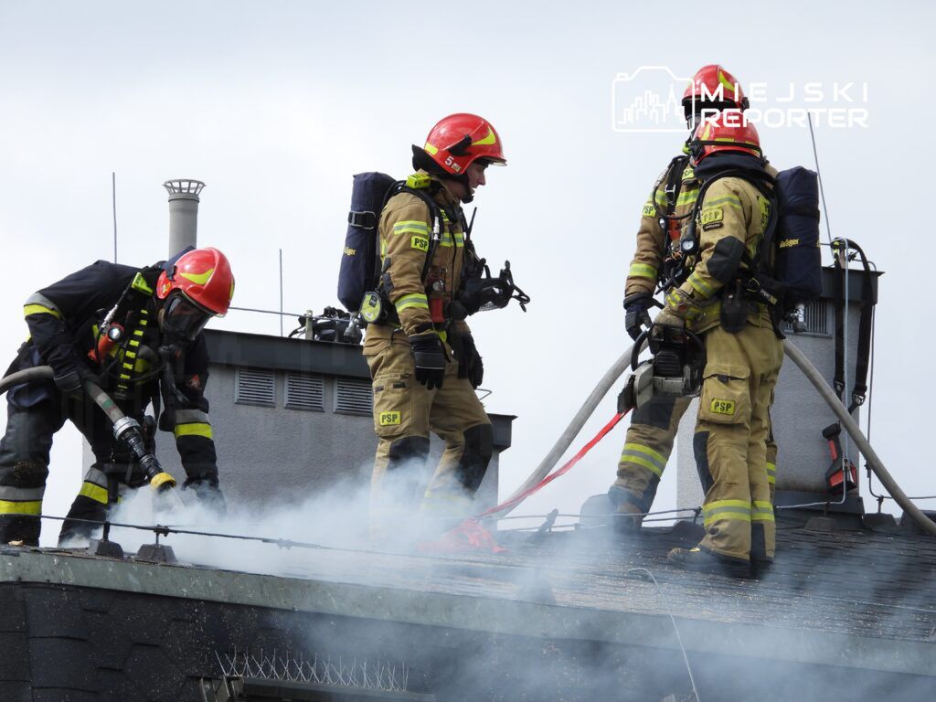 Grupa strażaków w hełmach i mundurach gaśniczych gasi dymiący dach budynku, używając węża pożarniczego.