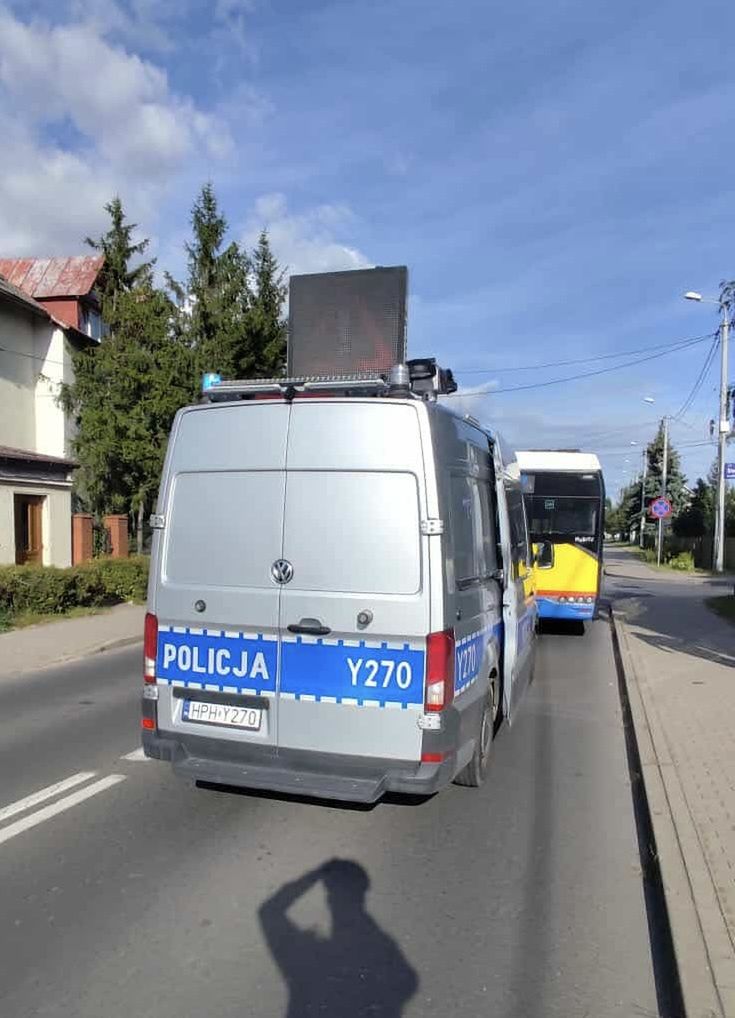 Policyjny samochód z oznaczeniem "POLICJA Y270" zatrzymany na ulicy, obok autobusu. W tle widoczne drzewa i budynki.