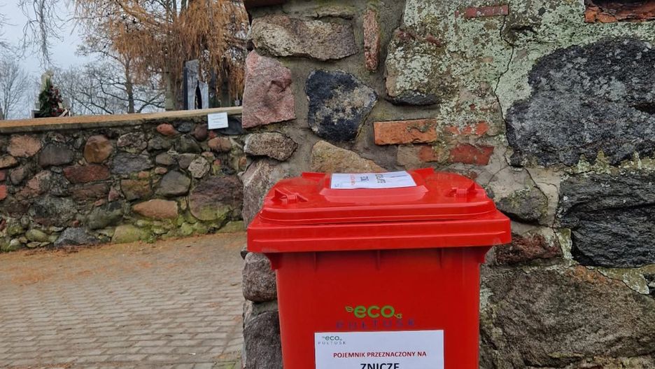Czerwony pojemnik na zużyte znicze elektryczne ustawiony przy wejściu na cmentarz w Pułtusku.