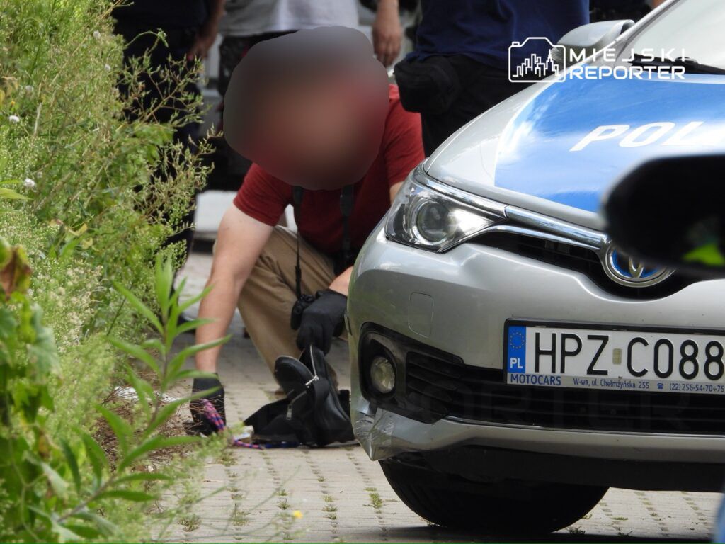 Funkcjonariusz policji przeszukuje teren obok radiowozu, w otoczeniu zieleni i chodnika.