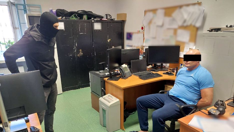 Mężczyzna w czarnej masce stoi obok siedzącego mężczyzny w niebieskiej koszulce, w biurze z komputerami.