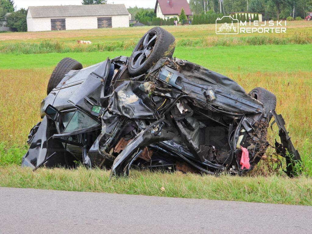 Uszkodzony samochód leży na dachu w polu, w tle widać stodołę oraz zieleń.
