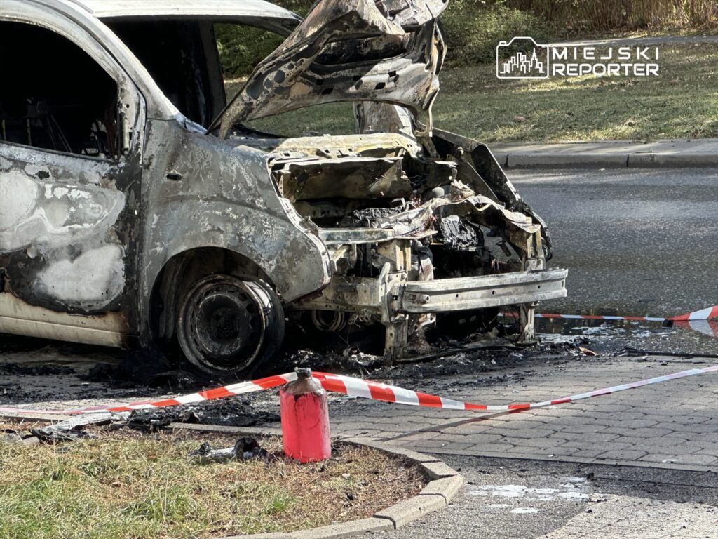 Spalony samochód osobowy z uszkodzoną karoserią stoi na chodniku, otoczony taśmą ostrzegawczą.