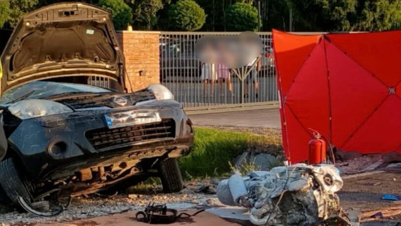 Zniszczony samochód leży na boku obok drogi, obok niego przewrócony silnik i czerwona parasolka ochronna.