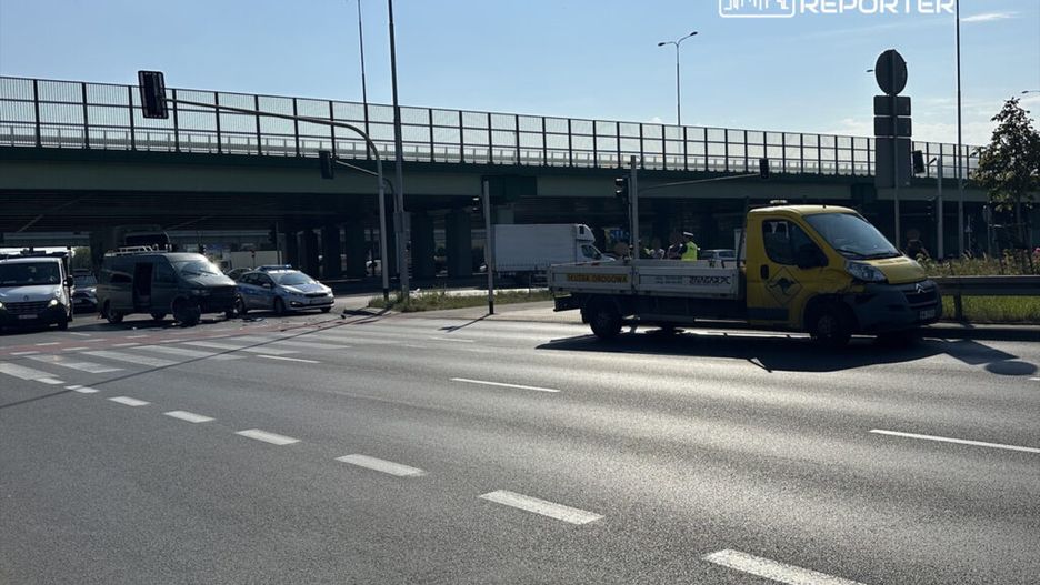 Na skrzyżowaniu stoi żółta ciężarówka z napisem "Służba Drogowa" obok uszkodzonego pojazdu, a w tle widać ruch uliczny.