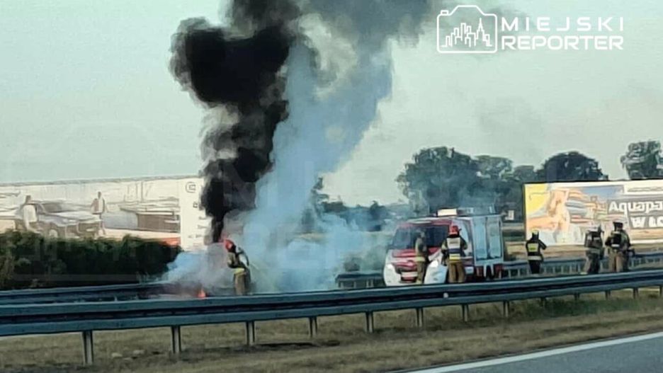 Strażacy gaszą pożar pojazdu na poboczu drogi, obok widoczna karetka i dym unoszący się w powietrzu.