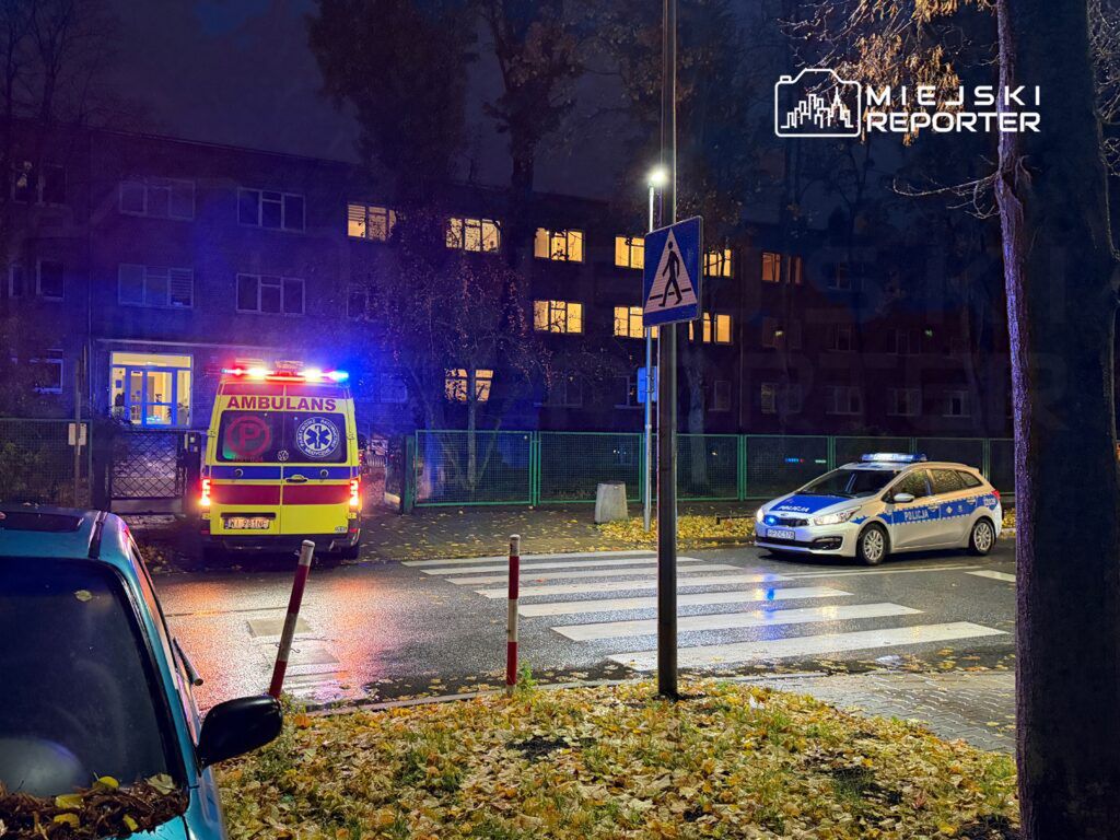 Karetka pogotowia z włączonymi sygnałami świetlnymi stoi przed budynkiem mieszkalnym, obok policyjny radiowóz.